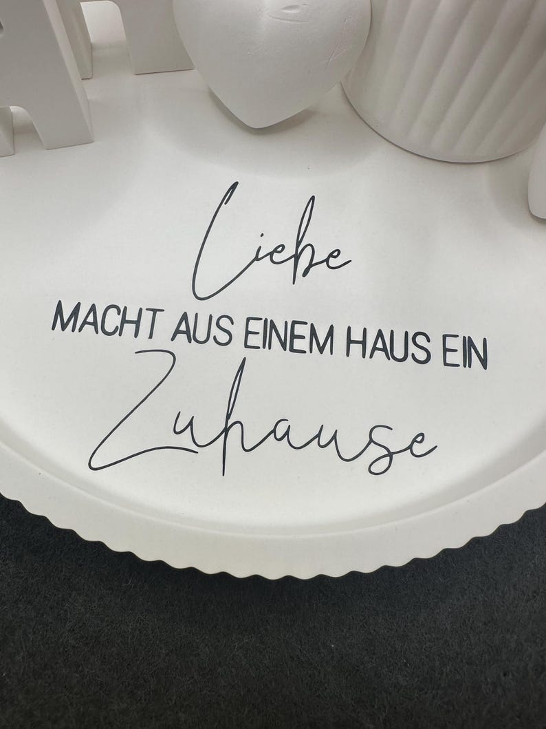 Puede incluir: Una bandeja blanca con borde festoneado con la frase alemana "Liebe macht aus einem Haus ein Zuhause" en escritura negra. Objetos decorativos blancos son parcialmente visibles en la bandeja.