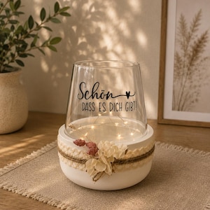 May include: A clear glass vase sits inside a white circular base decorated with a beige lace ribbon, twine, and small dried flowers. The glass features the black text "Sch&ouml;n DASS ES DICH GIBT!" with a heart symbol. A string of warm LED lights glows inside.