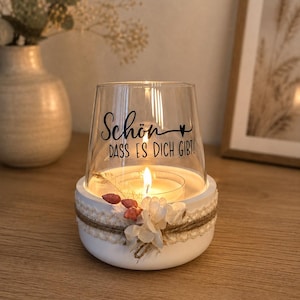 May include: A clear glass candle holder with a white circular base, decorated with a jute twine wrap, white lace trim, and small dried flowers. The glass features the black text "Sch&ouml;n DASS ES DICH GIBT!" with a small heart symbol.