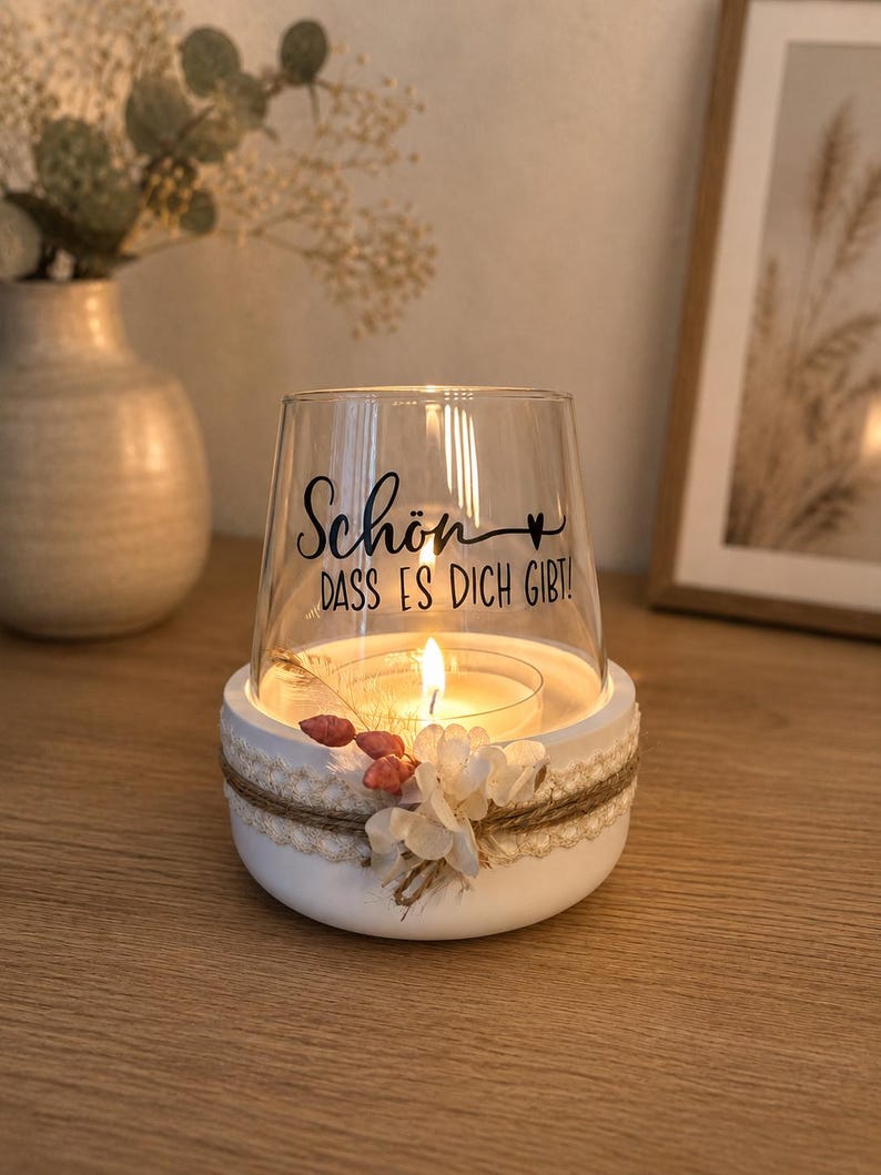 May include: A clear glass candle holder with a white circular base, decorated with a jute twine wrap, white lace trim, and small dried flowers. The glass features the black text "Sch&ouml;n DASS ES DICH GIBT!" with a small heart symbol.