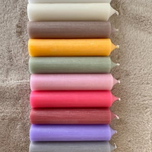 K&ouml;nnte beinhalten: Eine Sammlung von zehn farbigen Stabkerzen in verschiedenen Farbt&ouml;nen, darunter Wei&szlig;, Beige, Braun, Gelb, Gr&uuml;n, Rosa, Rot, Burgunder, Lila und Grau. Die Kerzen sind vertikal angeordnet und zeigen ihre zylindrische Form und ihre Spitzen.