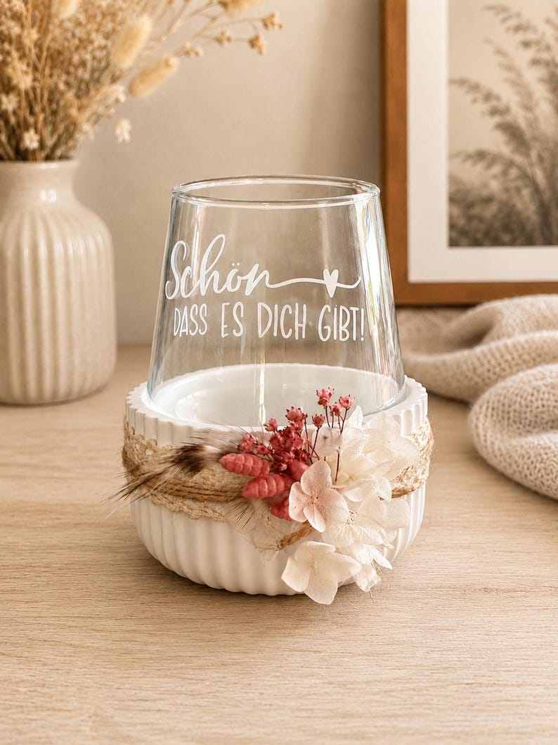May include: A clear glass tumbler featuring the white text "Sch&ouml;n DASS ES DICH GIBT!" with a small heart icon. The glass sits inside a white, ribbed ceramic bowl decorated with a jute twine wrap, dried pink flowers, and white hydrangea petals.