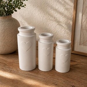 May include: Three off-white, cylindrical vases of varying sizes, each with a heart-shaped indentation. The vases have a rounded top and sit on a wooden surface. A large vase with greenery is in the background.