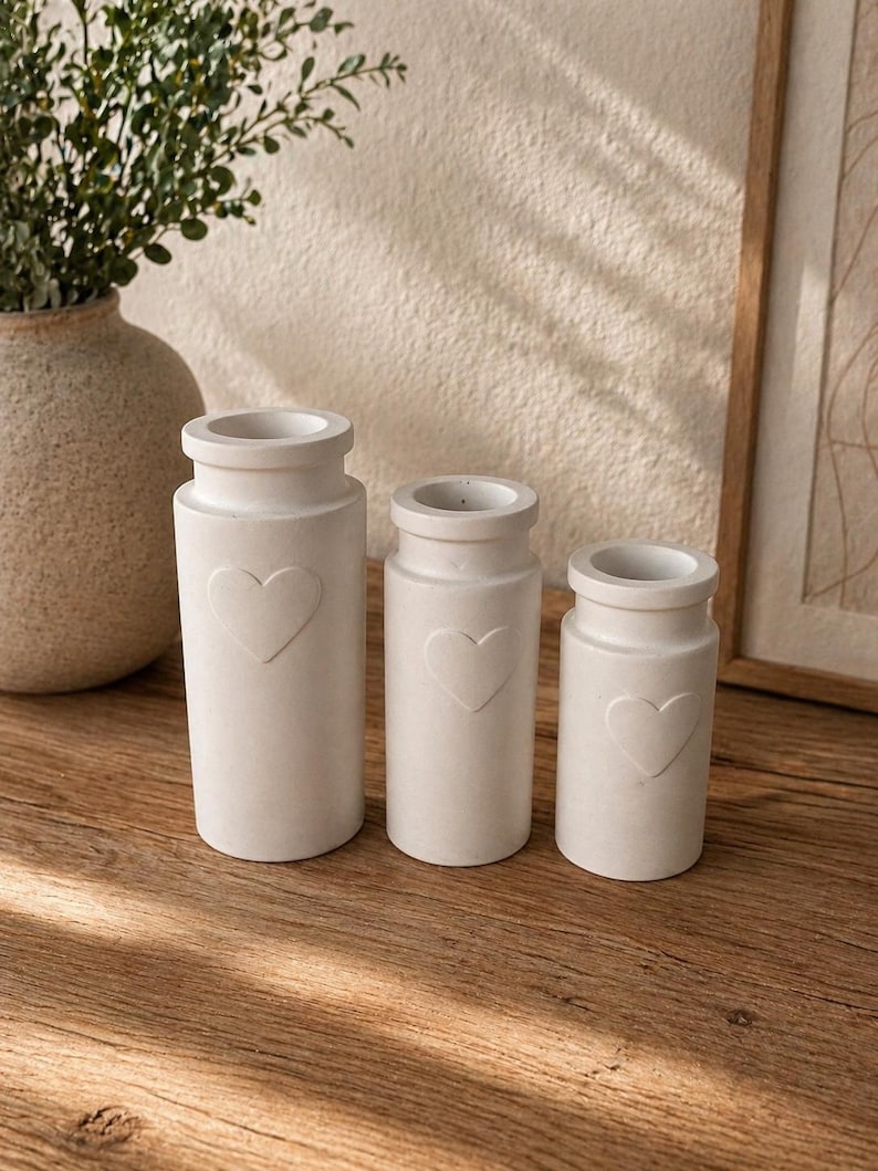 May include: Three off-white, cylindrical vases of varying sizes, each with a heart-shaped indentation. The vases have a rounded top and sit on a wooden surface. A large vase with greenery is in the background.