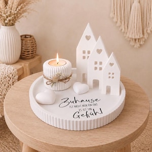 Puede incluir: Una bandeja de cer&aacute;mica blanca con una vela, casas decorativas y detalles en forma de coraz&oacute;n. La bandeja tiene el texto "Zuhause ist nicht nur ein Ort es ist ein Gef&uuml;hl" en escritura negra. Dispuesto sobre una peque&ntilde;a mesa de madera.