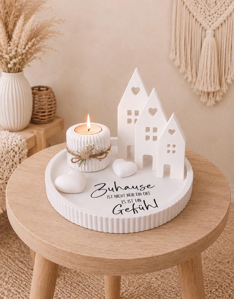 Puede incluir: Una bandeja de cer&aacute;mica blanca con una vela, casas decorativas y detalles en forma de coraz&oacute;n. La bandeja tiene el texto "Zuhause ist nicht nur ein Ort es ist ein Gef&uuml;hl" en escritura negra. Dispuesto sobre una peque&ntilde;a mesa de madera.