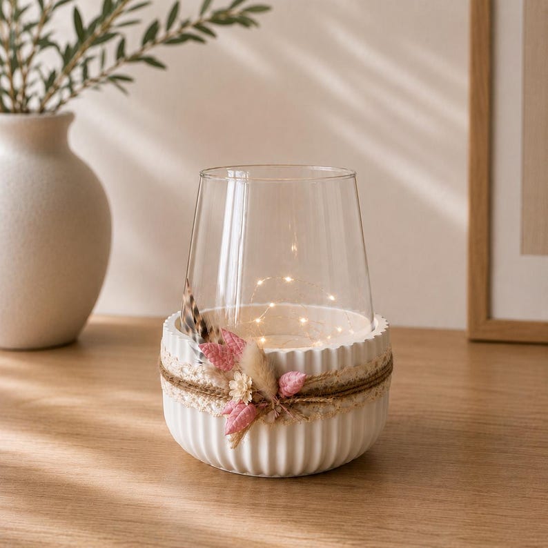 May include: A clear glass candle holder set inside a white, vertically ribbed ceramic bowl. The bowl is decorated with a band of beige lace, natural twine, and a cluster of small pink and cream-colored dried flowers and a single brown feather.