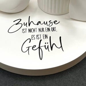 May include: A white, round decorative tray with the German text "Zuhause ist nicht nur ein Ort, es ist ein Gef&uuml;hl" in black script. The tray has a few white decorative objects on top, including a heart shape.