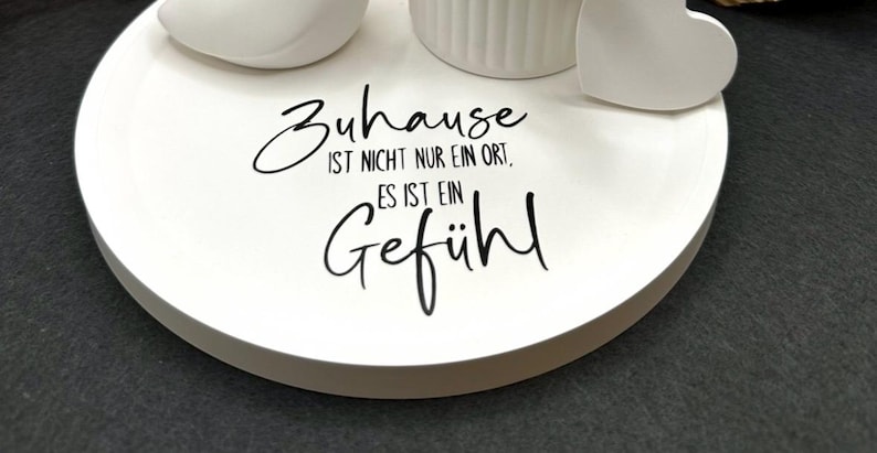 May include: A white, round decorative tray with the German text "Zuhause ist nicht nur ein Ort, es ist ein Gef&uuml;hl" in black script. The tray has a few white decorative objects on top, including a heart shape.