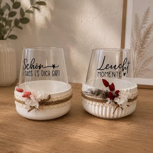May include: Two clear glass candle holders set in white ribbed ceramic bases. The left glass features the black text "Sch&ouml;n DASS ES DICH GIBT!" and the right glass features "Leucht MOMENTE". Both are decorated with twine, lace, and dried flowers.
