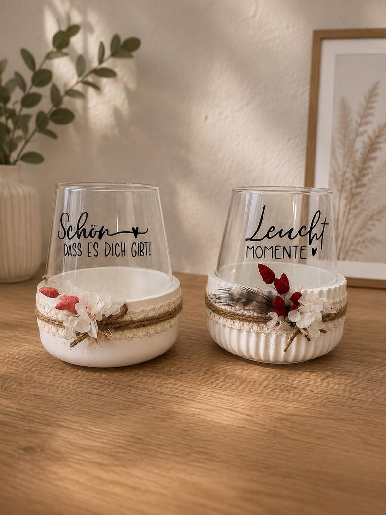 May include: Two clear glass candle holders set in white ribbed ceramic bases. The left glass features the black text "Sch&ouml;n DASS ES DICH GIBT!" and the right glass features "Leucht MOMENTE". Both are decorated with twine, lace, and dried flowers.