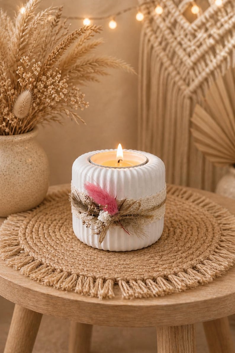 Puede incluir: Un portavelas de cer&aacute;mica blanca con una vela encendida. El portavelas est&aacute; decorado con una banda de encaje y flores secas. El portavelas se encuentra sobre un mantel individual redondo tejido sobre una mesa de madera.