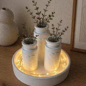 May include: A set of three white ceramic vases with heart-shaped cutouts, each holding greenery, arranged on a round, white tray. The tray is illuminated by a string of warm white fairy lights, creating a cozy, decorative display.