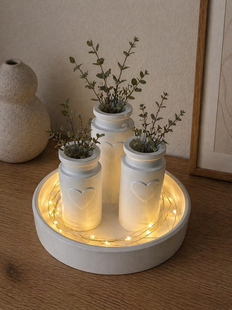 May include: A set of three white ceramic vases with heart-shaped cutouts, each holding greenery, arranged on a round, white tray. The tray is illuminated by a string of warm white fairy lights, creating a cozy, decorative display.