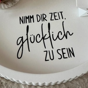 Puede incluir: Bandeja decorativa redonda blanca con borde festoneado. La bandeja presenta el texto alem&aacute;n "Nimm dir Zeit. gl&uuml;cklich ZU SEIN" en escritura negra. Se ven una peque&ntilde;a estrella blanca y detalles florales.