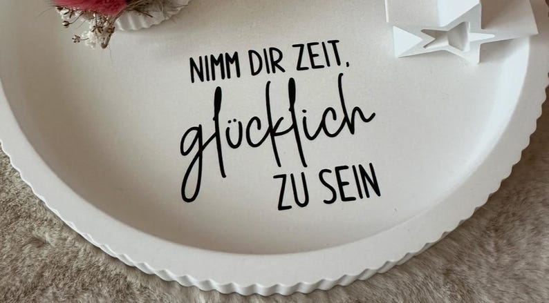 Puede incluir: Bandeja decorativa redonda blanca con borde festoneado. La bandeja presenta el texto alem&aacute;n "Nimm dir Zeit. gl&uuml;cklich ZU SEIN" en escritura negra. Se ven una peque&ntilde;a estrella blanca y detalles florales.