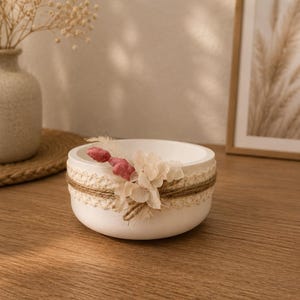 May include: A round white bowl decorated with a band of cream-colored lace and natural twine. The exterior is adorned with a small cluster of dried flowers, including pale pink and off-white petals, attached to the side of the bowl.