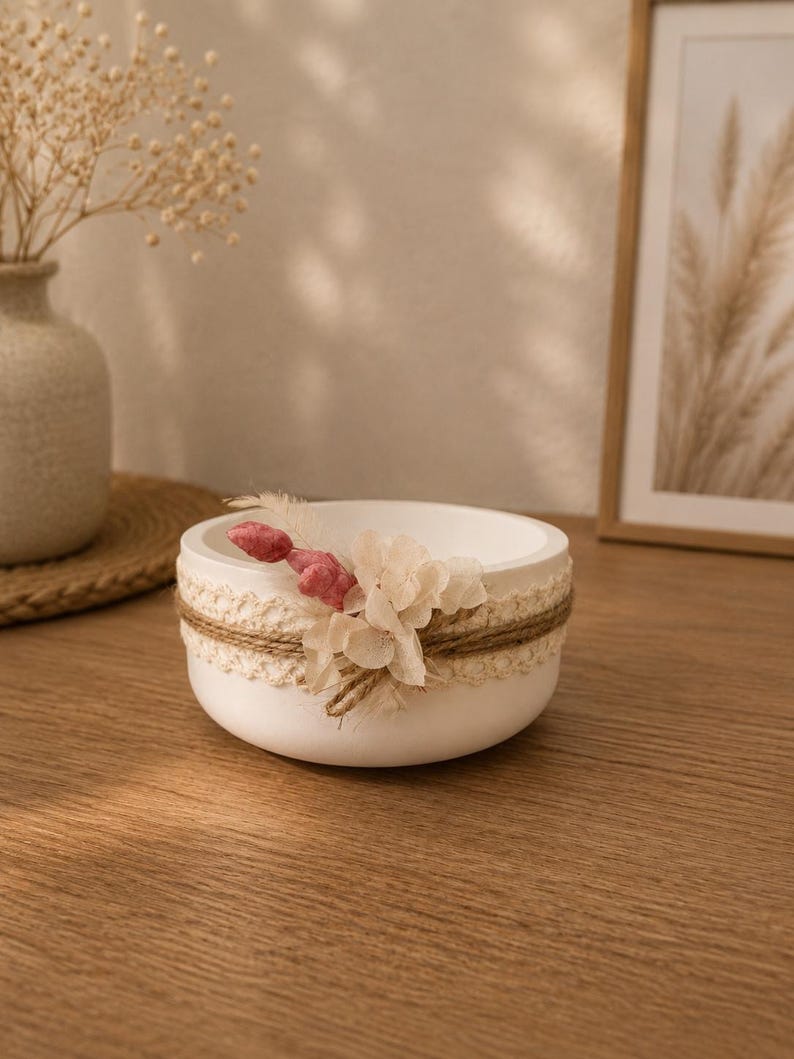 May include: A round white bowl decorated with a band of cream-colored lace and natural twine. The exterior is adorned with a small cluster of dried flowers, including pale pink and off-white petals, attached to the side of the bowl.
