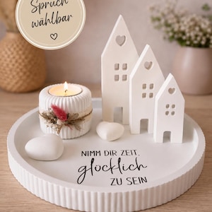 May include: A white decorative tray with a candle holder, small ceramic houses, and heart-shaped stones. The candle holder has a lit candle and is decorated with dried flowers. The tray has the text "NIMM DIR ZEIT. gl&uuml;cklich ZU SEIN."