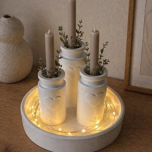 May include: A decorative centerpiece featuring three white ceramic candle holders with heart cutouts, each holding a taupe candle and greenery. The holders sit on a round, white tray with string lights, creating a warm, inviting ambiance.