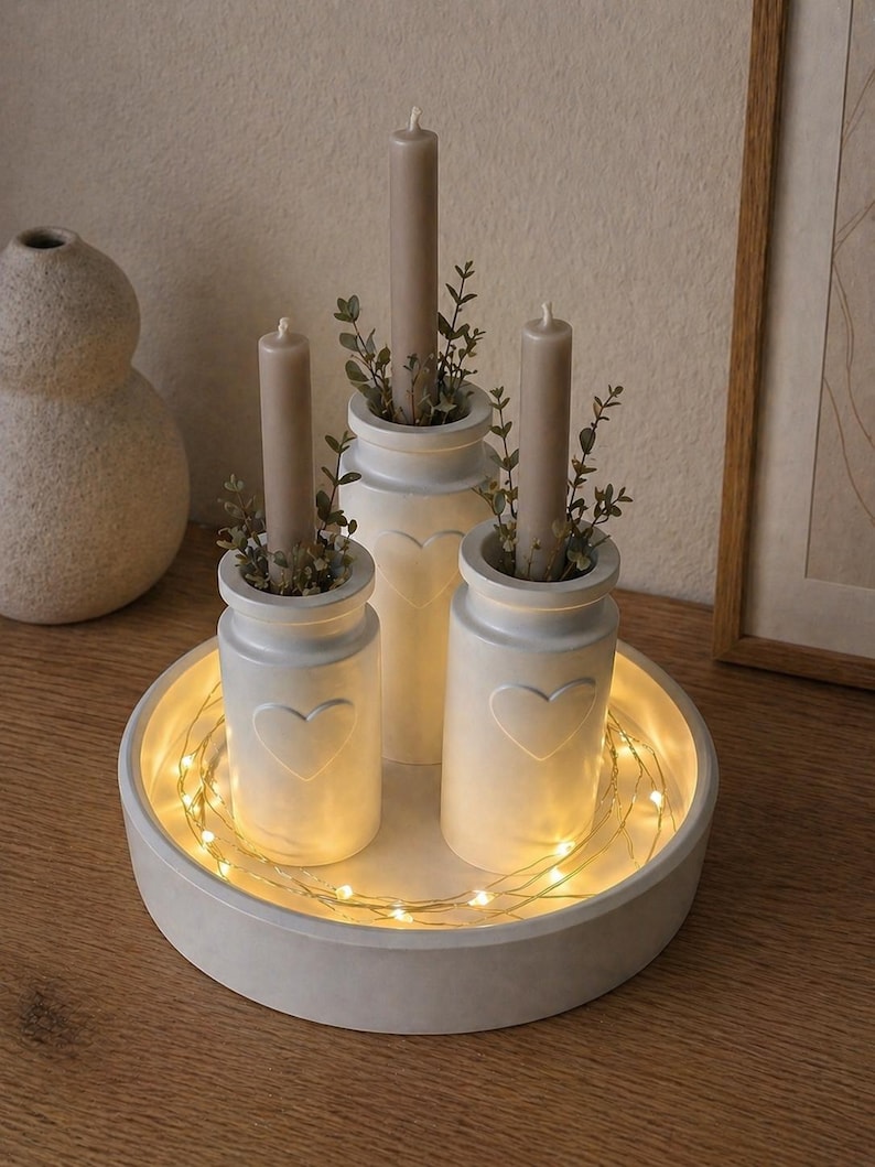 May include: A decorative centerpiece featuring three white ceramic candle holders with heart cutouts, each holding a taupe candle and greenery. The holders sit on a round, white tray with string lights, creating a warm, inviting ambiance.