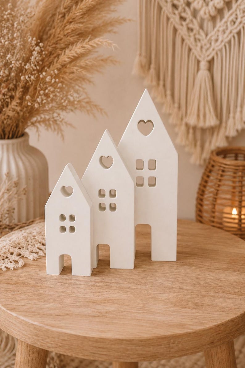 Puede incluir: Tres figuras de casas de cer&aacute;mica blanca con recortes en forma de coraz&oacute;n y ventanas cuadradas. Las casas son de diferentes tama&ntilde;os y est&aacute;n sobre una mesa de madera. El fondo incluye plantas secas y un tapiz de pared tejido.