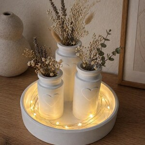 May include: A white, round tray with a recessed center holds three white vases, each with a heart design. The vases contain dried flowers and are illuminated by string lights. The arrangement sits on a wooden surface.
