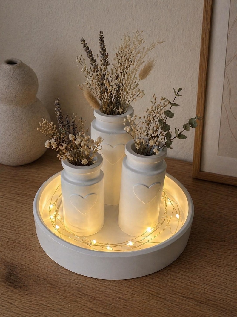 May include: A white, round tray with a recessed center holds three white vases, each with a heart design. The vases contain dried flowers and are illuminated by string lights. The arrangement sits on a wooden surface.