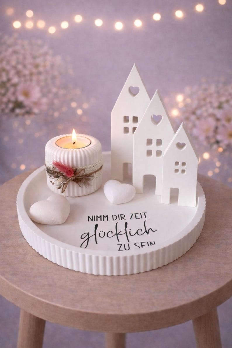 May include: A white ceramic tray with a candle holder, three house-shaped decorations, and two heart-shaped decorations. The candle holder has a lit candle and is decorated with lace and feathers. The tray has the text "Nimm dir Zeit, gl&uuml;cklich zu sein."