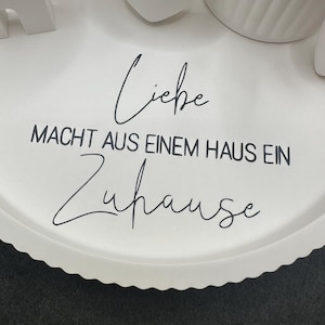 May include: A white, scalloped-edge tray with the German phrase "Liebe macht aus einem Haus ein Zuhause" in black script. Decorative white objects are partially visible on the tray.