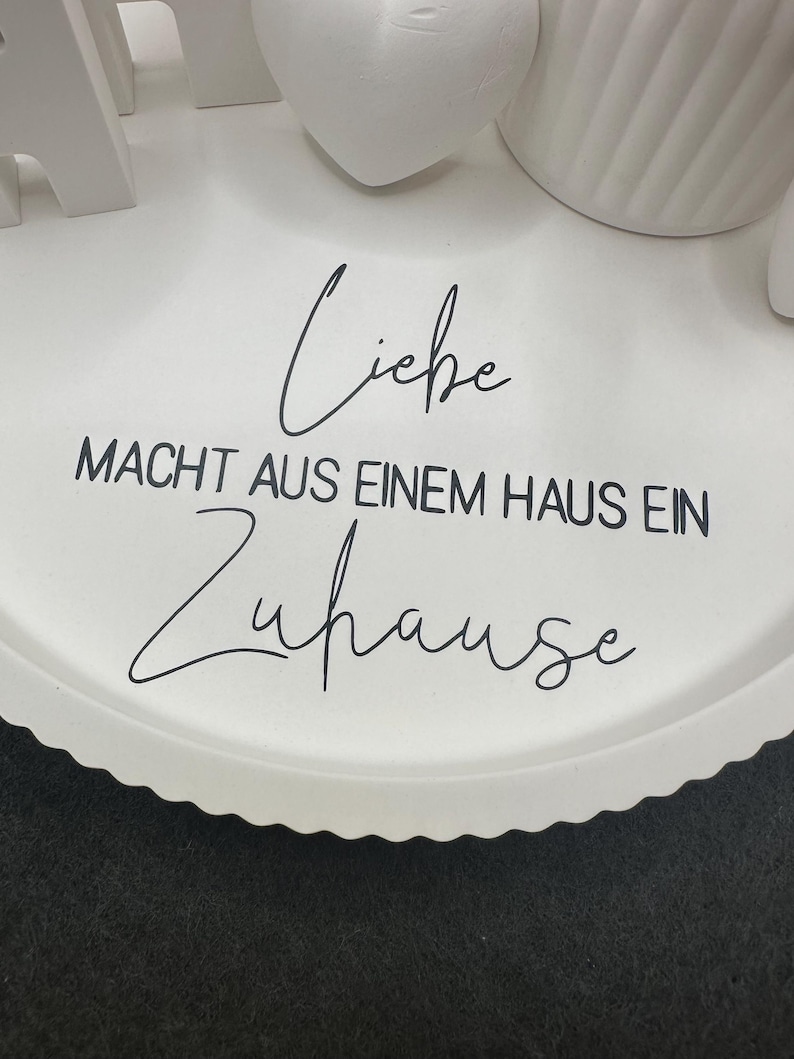 May include: A white, scalloped-edge tray with the German phrase "Liebe macht aus einem Haus ein Zuhause" in black script. Decorative white objects are partially visible on the tray.
