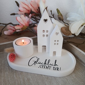 Könnte beinhalten: Ein weißer Kerzenhalter mit einer kleinen brennenden Kerze, zwei hausförmigen Dekorationen und dem Text "Glücklich. STEHT DIR!" in schwarzer Schrift. Rosa und weiße Blumen sind im Hintergrund.