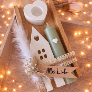 K&ouml;nnte beinhalten: Ein Holztablett mit einem wei&szlig;en herzf&ouml;rmigen Kerzenhalter, einem hausf&ouml;rmigen Ornament und einer gr&uuml;nen Kerze mit einem Herz. Ein Banner mit dem Text "Alles Liebe" ist enthalten. Die Anordnung ist mit Federn und Blumen dekoriert.