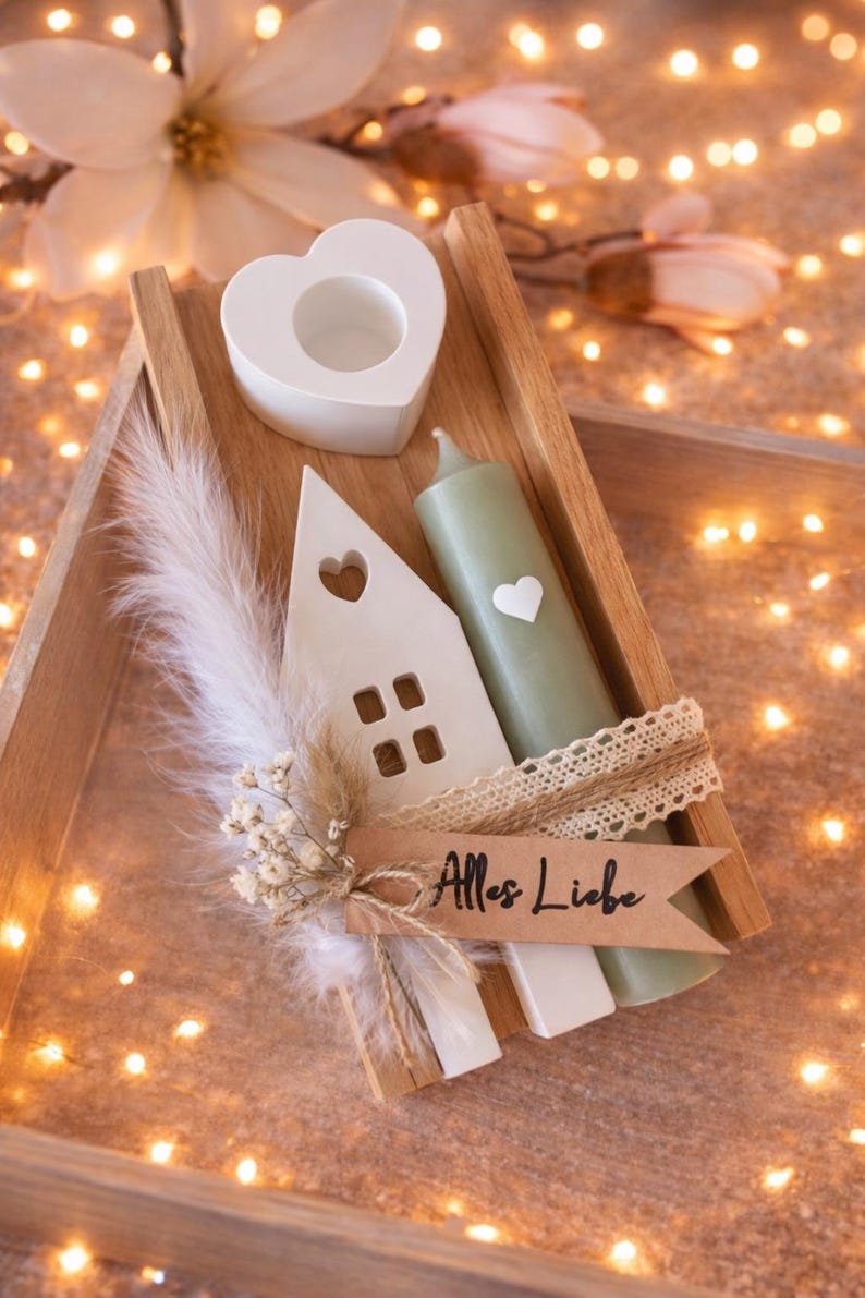 K&ouml;nnte beinhalten: Ein Holztablett mit einem wei&szlig;en herzf&ouml;rmigen Kerzenhalter, einem hausf&ouml;rmigen Ornament und einer gr&uuml;nen Kerze mit einem Herz. Ein Banner mit dem Text "Alles Liebe" ist enthalten. Die Anordnung ist mit Federn und Blumen dekoriert.