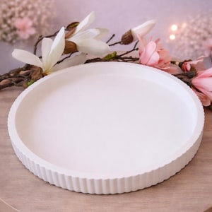 May include: A white, circular serving tray with a fluted edge, displayed on a wooden table. The tray has a smooth surface. Decorative flowers and soft lighting are in the background.
