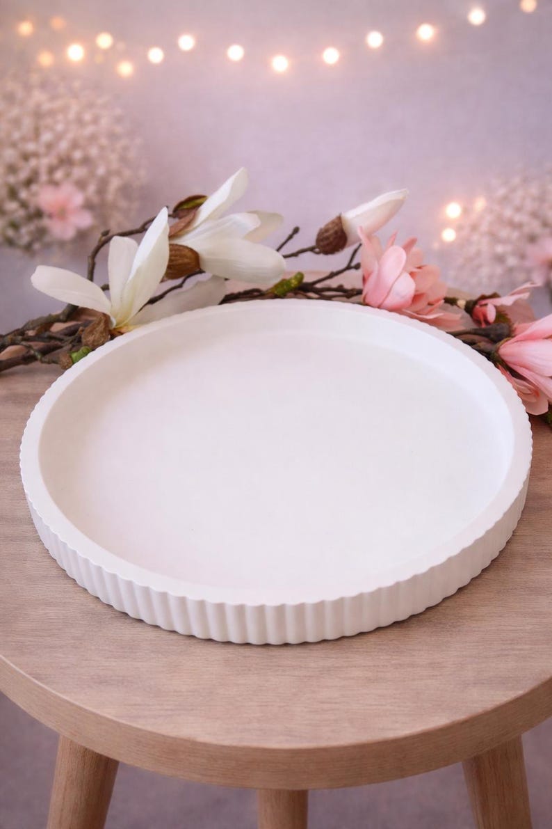 May include: A white, circular serving tray with a fluted edge, displayed on a wooden table. The tray has a smooth surface. Decorative flowers and soft lighting are in the background.