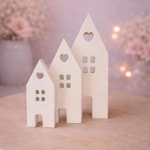 May include: Three off-white ceramic house ornaments of different sizes. Each house features a heart-shaped cutout and square window cutouts. The houses are displayed on a light brown wooden table, with a soft-focus background of flowers and lights.
