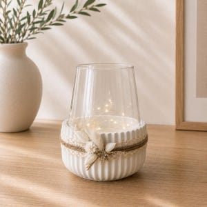 May include: A clear glass hurricane candle holder set inside a white fluted ceramic base. The base is wrapped with natural jute twine and decorated with a cluster of dried white flowers and grasses. A string of warm white fairy lights glows inside.