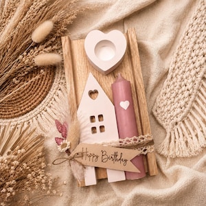 K&ouml;nnte beinhalten: Eine dekorative Holzkomposition mit einem herzf&ouml;rmigen Kerzenhalter, einem hausf&ouml;rmigen Ornament mit Herz-Ausschnitten und einer rosa Kerze mit einem Herz. Ein Banner mit "Happy Birthday" ist angebracht. Die Anordnung befindet sich auf einem Holztablett.