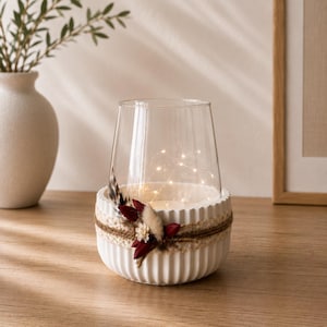 May include: A clear glass candle holder sits inside a white, vertically ribbed ceramic bowl. The bowl is wrapped with a natural jute twine band decorated with small dried flowers in shades of deep red and cream, along with a single brown feather.