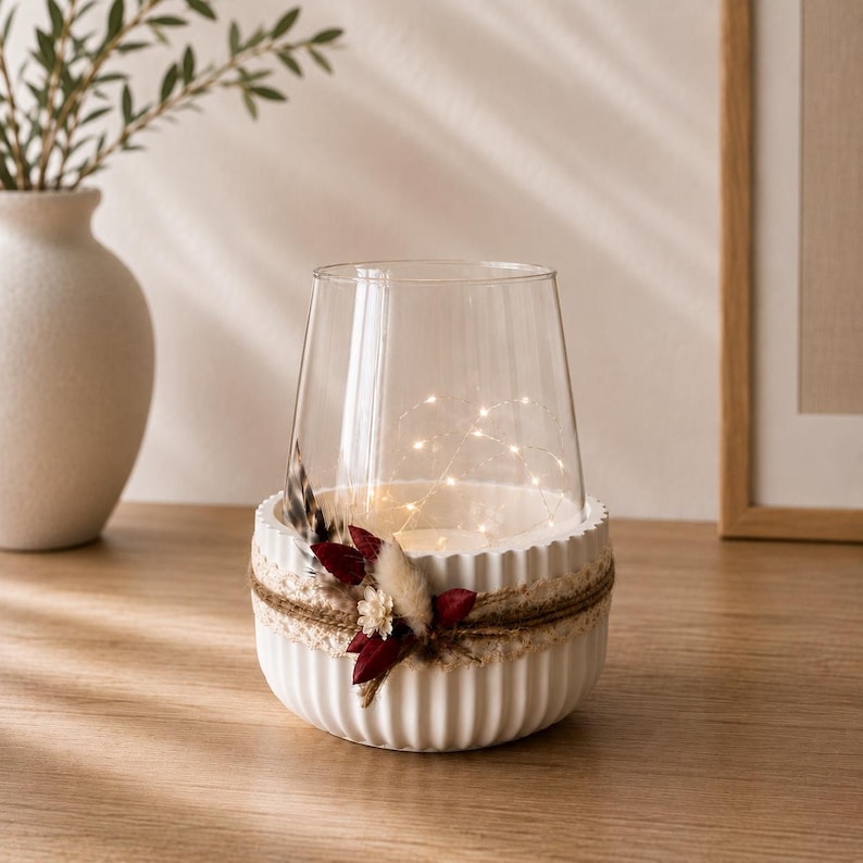 May include: A clear glass candle holder sits inside a white, vertically ribbed ceramic bowl. The bowl is wrapped with a natural jute twine band decorated with small dried flowers in shades of deep red and cream, along with a single brown feather.