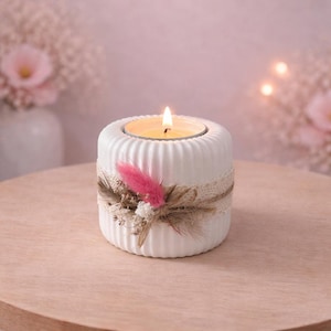 May include: A white, ribbed ceramic candle holder with a lit candle. The holder is decorated with a lace band, dried flowers, and a pink feather. The candle holder sits on a wooden table.