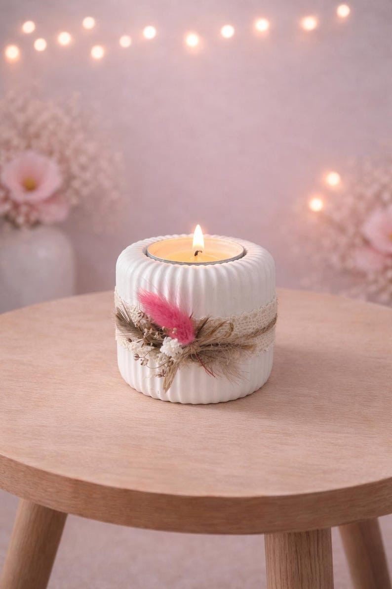 May include: A white, ribbed ceramic candle holder with a lit candle. The holder is decorated with a lace band, dried flowers, and a pink feather. The candle holder sits on a wooden table.