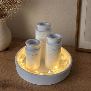 May include: A white, round tray holds three small, white, cylindrical vases with heart-shaped cutouts. The vases are lit from within, and the tray is encircled by a string of warm white fairy lights. The arrangement sits on a wooden surface.