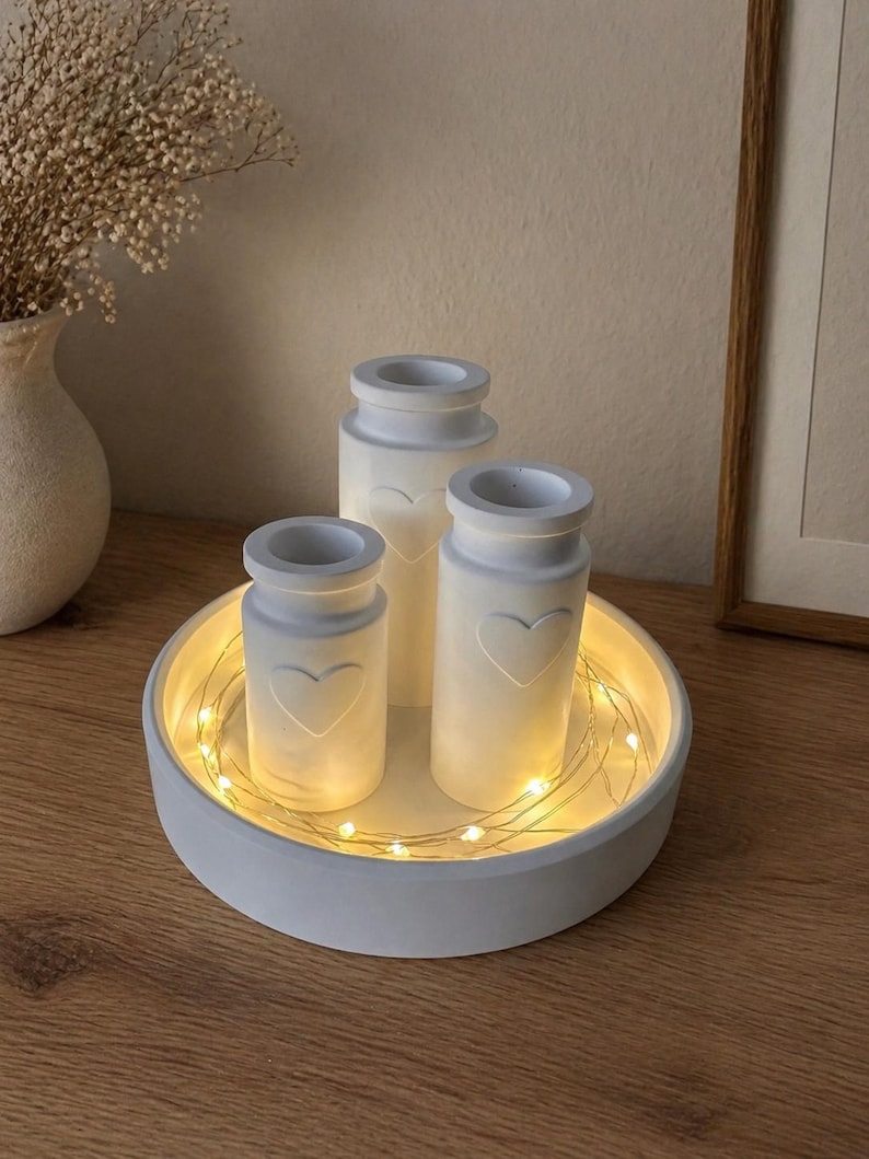 May include: A white, round tray holds three small, white, cylindrical vases with heart-shaped cutouts. The vases are lit from within, and the tray is encircled by a string of warm white fairy lights. The arrangement sits on a wooden surface.