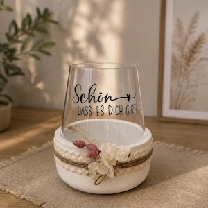 May include: A clear glass vessel sits inside a white circular base. The glass features black text that reads "Sch&ouml;n, dass es dich gibt!" with a small heart symbol. The white base is decorated with a beige lace ribbon, twine, and dried flowers.