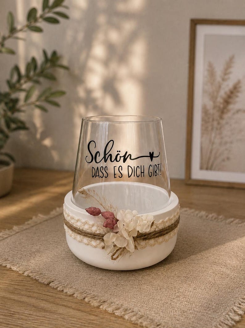 May include: A clear glass vessel sits inside a white circular base. The glass features black text that reads "Sch&ouml;n, dass es dich gibt!" with a small heart symbol. The white base is decorated with a beige lace ribbon, twine, and dried flowers.