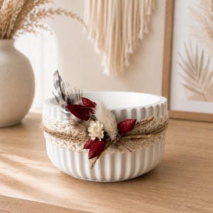 May include: A white, round, fluted bowl decorated with a band of beige lace and twine. The arrangement features dried elements including a spotted feather, small cream-colored flowers, and deep red dried leaves attached to the front.