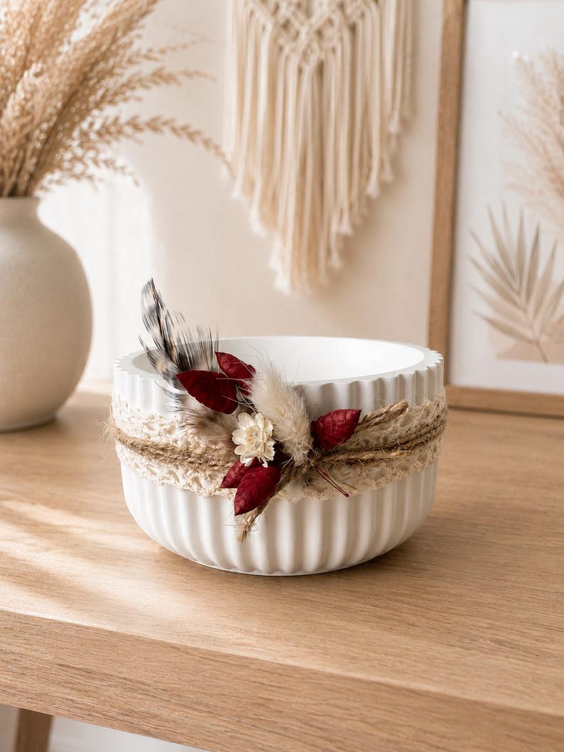 May include: A white, round, fluted bowl decorated with a band of beige lace and twine. The arrangement features dried elements including a spotted feather, small cream-colored flowers, and deep red dried leaves attached to the front.