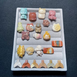 May include: A collection of small, carved gemstone figurines in various shapes, including animals and abstract forms. The figurines are displayed in a gray tray, with colors ranging from pastel blues and pinks to earthy browns and yellows. The word "Front" is visible in yellow.